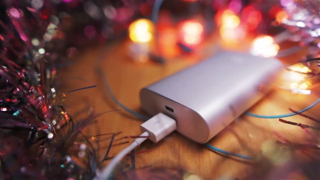 Silver powerbank external battery for fast charging smartphone or tablet pc lying on the table. surrounded by flashing garland lights.