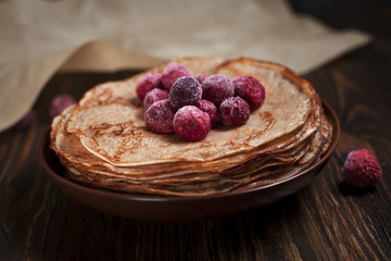 pancakes with frozen berries pile in a brown plate on a dark wooden table on a dark gray background