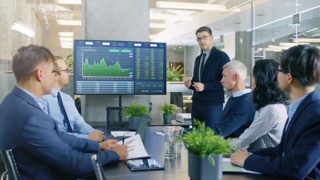 Young Stock Trader Shows to the Executive Managers Cryptocurrency and Trade Market Correlation Pointing at the Wall TV. Shot on RED EPIC-W 8K Helium Cinema Camera.