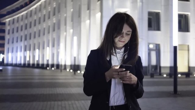 Attractive young dark haired woman wearing a suit is looking at her cell phone screen and walking in a night city. Tracking real time establishing shot