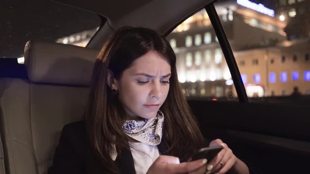 Attractive young businesswoman is sitting in a backseat of her car and frowning while looking at her smartphone. Locked down real time medium shot