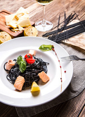 Pasta with cuttlefish ink, salmon and cherry tomatoes in a white plate on a dark wooden background top view