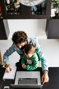 Man With Kid Writing Notes At Desk With Laptop.