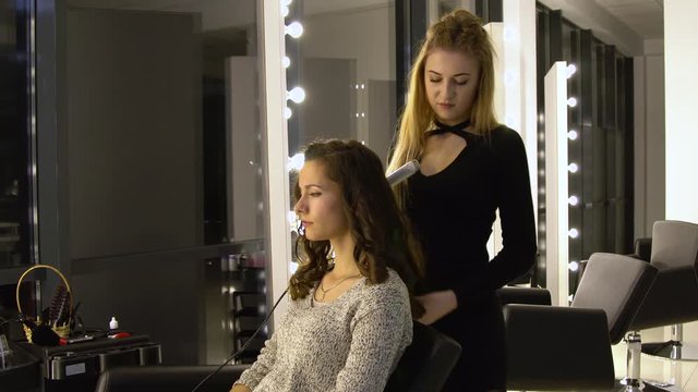 Young beautiful girl in beauty salon. Hairdresser-stylist makes the girl's hair. The hairdresser curls the hair with a styler and stacks them.