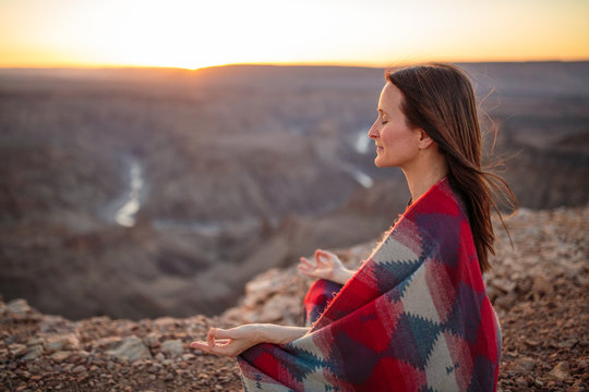 Beautiful woman wrapped in kaftan blanket meditating at sunset overlooking a canyon
