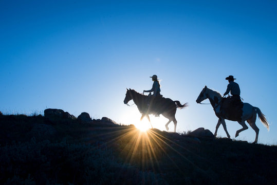 Silhouetted cowboy and cowgirl on horseback, with a blue sky and setting sun at horiozn