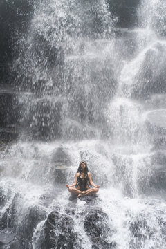 Young woman wearing swimsuit posing in lotus pose under waterfall