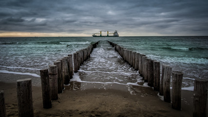 Fototapeta premium Big ship crossing the wooden pier during cloudy weather at the beach in Vlissingen, Zeeland, Holland, Netherlands