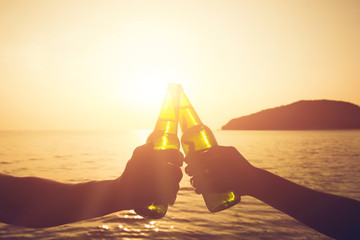 Couple hands holding beer bottles and clanging, celebrating on holiday at the beach in sunset