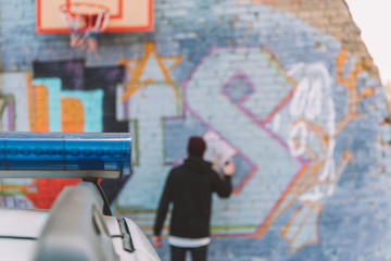 back view of vandal painting graffiti on wall, police car on foreground