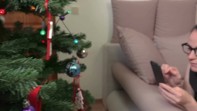 Camera focusing on Christmas decoration on Christmas tree then on woman who relaxing on sofa