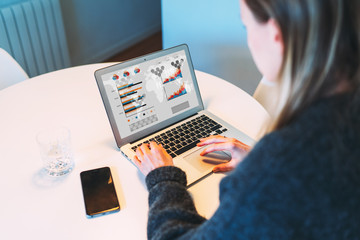 Young businesswoman is sitting at table and uses laptop with graphs, charts, diagrams on screen. Girl working online, online marketing education, training, webinar. On table smartphone. Rear view.