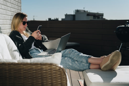Sunny day. Young businesswoman is sitting on white couch on terrace, using laptop and smartphone. Hipster girl working, blogging, learning online. Distance work.Online marketing,education, e-learning.
