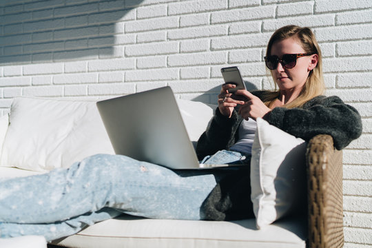 Sunny day. Young businesswoman is sitting on white couch on terrace, using laptop and smartphone. Hipster girl working, blogging, learning online. Distance work.Online marketing,education, e-learning.
