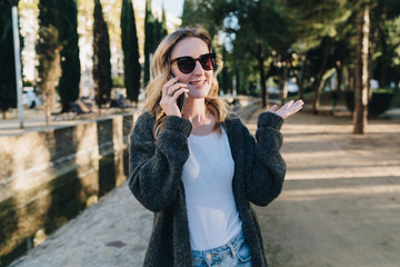 Summer evening. A young attractive woman in sunglasses stands in the park and talks happily on her cell phone. Telephone conversations. A girl is calling a friend on the phone. Lifestyle.