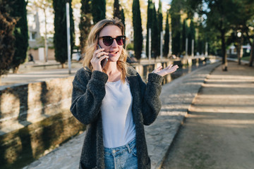 Fototapeta premium Summer evening. A young attractive woman in sunglasses stands in the park and talks happily on her cell phone. Telephone conversations. A girl is calling a friend on the phone. Lifestyle.