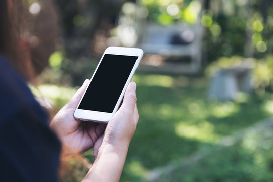 Mockup image of a woman holding and using white smart phone with blank black screen at outdoor and blur green nature background