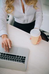 cropped image of businesswoman holding disposable coffee cup and using laptop