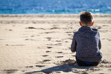 Sad young boy sitting on the beach, looking at sea and thinking
