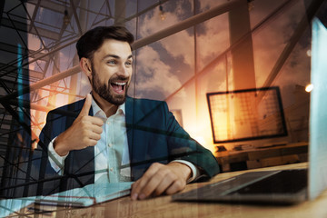 Fototapeta na wymiar Positive mood. Cheerful young businessman sitting at the office table and showing thumbs up to the web camera while having a video call with his partners