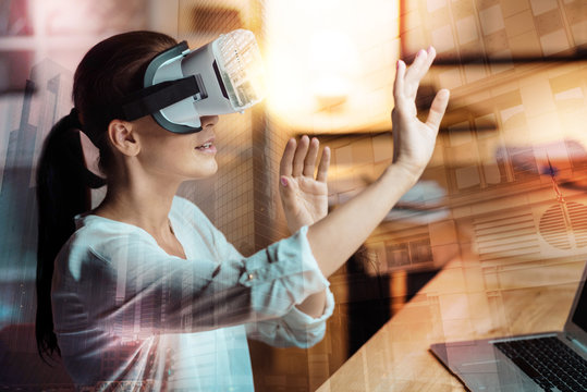 Brand New Technology. Pleasant Young Woman Sitting At The Table In The Office And Testing A New VR Headset, Stretching Out Her Hands As If Being Ready To Touch Something