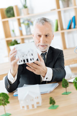 Nice work. Clever experienced worker of a real estate agency sitting in his office with a wonderful miniature of a house in his hands and looking attentive while thinking about it