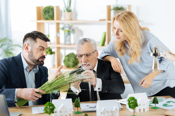 Interesting material. Emotional enthusiastic real estate agents looking curious while touching a bunch of synthetic grass and discussing its usage near one of their gorgeous houses