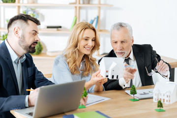 Amazing work. Smart interested aged real estate agent looking surprised and pointing at the lovely miniature house while his colleagues sitting by his side and looking attentively at it