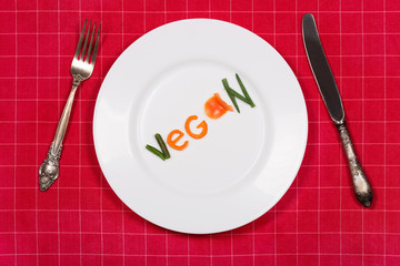 White plate with word vegan made of pieces of vegetables on red tablecloth background. Vintage fork...