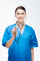 Stethoscope usage. Pretty joyful female doctor gazing  straight while carrying  stethoscope and trying to hear