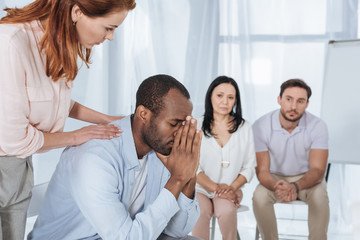 middle aged people supporting upset african american man during anonymous group therapy