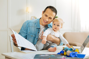 Obraz na płótnie Canvas New activity. Adorable little calm child feeling glad while sitting on the table next to the modern laptop and smiling while a loving kind smart father sitting near and working