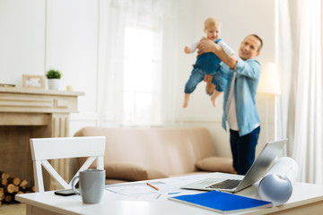 Favorite game. Cheerful kind attentive father having fun with his cute little baby after coming home from work and leaving all the important tasks on the table