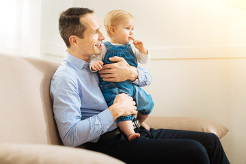 Watching film. Cheerful calm relaxed father sitting on the sofa and holding a little child while watching an interesting film together