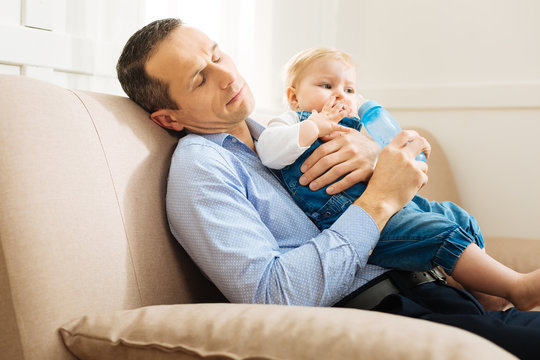 Sleepy. Exhausted Young Attentive Father Falling Asleep While Sitting On A Soft Comfortable Sofa With A Bottle Of Water In His Hand And Holding A Little Baby