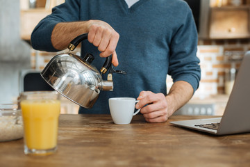 Making tea. Delighted man standing near table and raising right hand while pouring down water