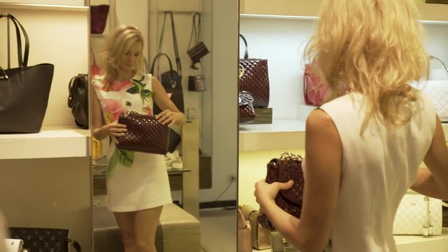The blonde tries on and chooses a ladies handbag standing by the mirror in the clothing shop. Shopping.