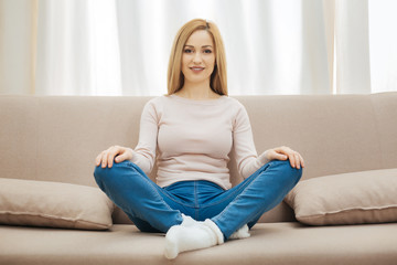 Relaxing. Happy young slim blond woman smiling and wearing jeans and a sweater and sitting on the couch