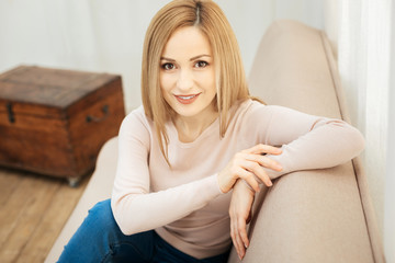 Over the moon. Pretty joyful young slim blond woman smiling and wearing jeans and a beige sweater and sitting on the couch in the room