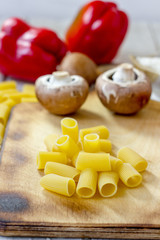 Dry rigatoni pasta on wooden kitchen board with vegetables.