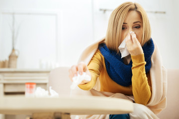 Blocked nose. Ill young blond woman blowing her nose while having a blanket on her shoulders while sitting on the couch and putting used napkins on the table in front of her