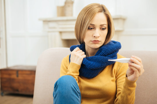 Feeling bad. Beautiful young blond woman feeling down in the mouth and holding a thermometer and having a blue warm scarf on while sitting on the sofa