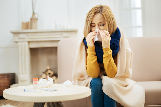 Under the weather. Ill young blond woman feeling bad and blowing her nose while having a blanket on her shoulders and sitting on the couch with her eyes closed and table with pills in front of her