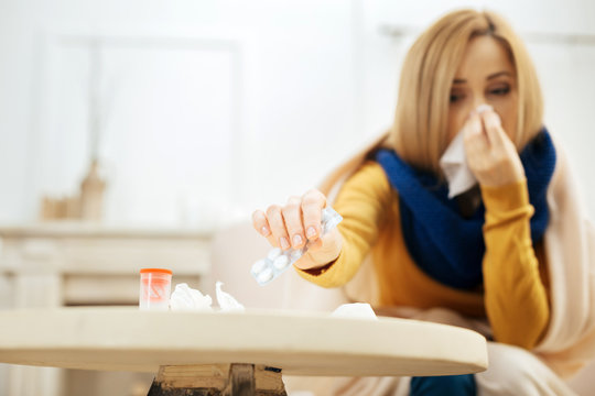 Medications. Sick young blond woman blowing her nose while having a blanket on her shoulders and sitting on the couch and taking the pills from the table in front of her