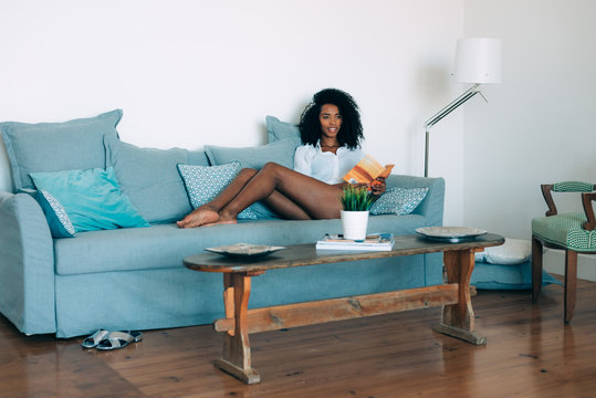 Beautiful young black woman seating in the sofa reading a book.