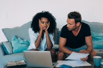 Young interracial couple in the couch stressed with financial problems doing calculations with paper work.