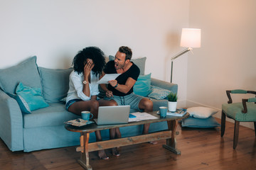 Young interracial couple in the couch stressed with financial problems doing calculations with paper work.