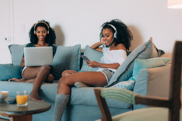 Happy young two black women sitting in the couch on the computer and mobile phone with headphones .