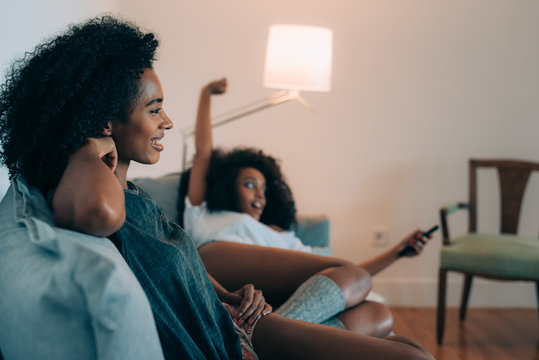 Happy Young Two Black Women Lying Down In The Couch Watching Tv  .