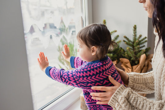 Close-up Portrait Of Mother With Daughter Sitting By The Window. On Christmas, Waiting For A Miracle. Beautiful Caucasian Woman Holding A Child Looking Out The Window With Hands On It.